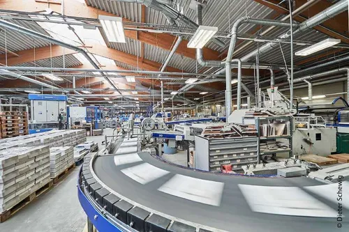 Eine große Fabrik mit Förderbändern, Industriemaschinen und Lagerboxen. Die Decke hat Metallträger und Leuchtstofflampen. Arbeiter sind anwesend.