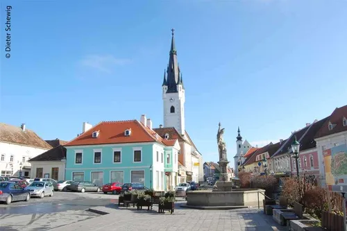 Ein Stadtplatz mit einem Brunnen, einer Statue und farbenfrohen Gebäuden. Im Hintergrund ist ein Kirchturm zu sehen. Autos sind am Straßenrand geparkt.