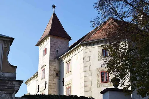 Ein Turm aus Stein mit einem spitzen Dach ist Teil eines größeren Gebäudes. Das Gebäude hat ein rotes Ziegeldach, mehrere Fenster und dekorative Elemente.