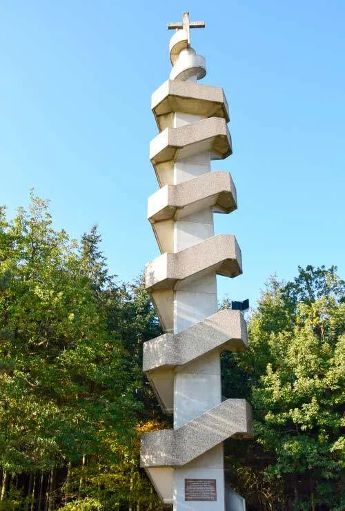 Ein Betonmonument mit einem Kreuz auf der Spitze wird von einem Wald unter klarem Himmel umgeben.