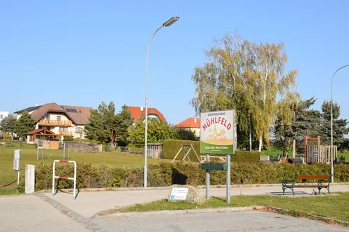 Ein Schild steht im Park und zeigt 'Dorferneuerunng Mühlfeld'. Es gibt Häuser, Bäume und ein Spielplatz.