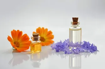 Zwei kleine Flaschen mit Korkverschlüssen, gefüllt mit ätherischen Ölen, neben Blumen auf einer weißen Oberfläche platziert. Eine orangefarbene Margerite und violette Veilchen sind sichtbar.