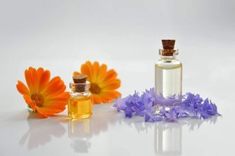 Zwei kleine Flaschen mit Korkverschlüssen, gefüllt mit ätherischen Ölen, neben Blumen auf einer weißen Oberfläche platziert. Eine orangefarbene Margerite und violette Veilchen sind sichtbar.