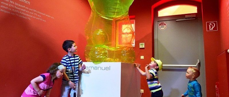 Vier Kinder stehen in einem Raum und betrachten eine große, bunte Skulptur. Hinter ihnen befindet sich eine rote Wand mit einem weißen Schild, und rechts ist eine Tür.
