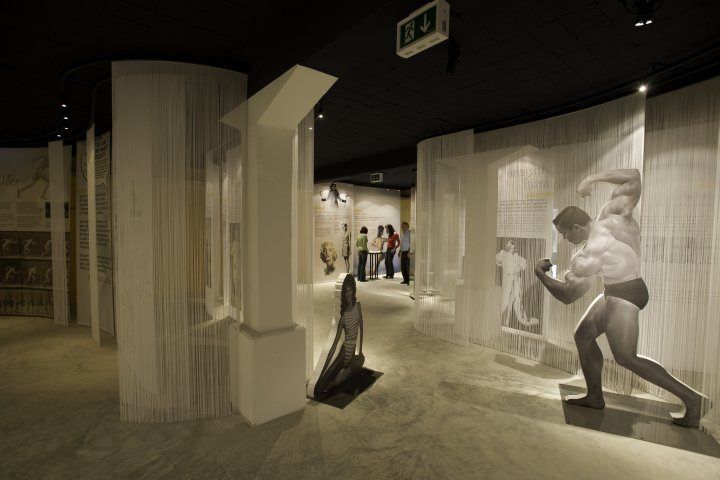 Eine schwach beleuchtete Indoor-Ausstellung mit großen Wandinstallationen und Skulpturen, einschließlich einer sitzenden Frau und eines muskulösen Mannes in Bewegung. Besucher sind anwesend.