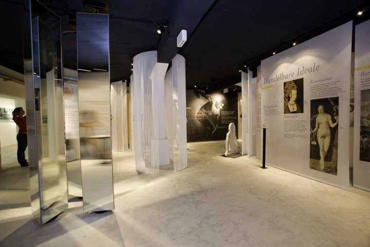 Ein Ausstellungsraum mit einer Person, die steht, und großen weißen Säulen. Die Wände haben Displays mit Gemälden und der Boden ist glänzend und sauber.
