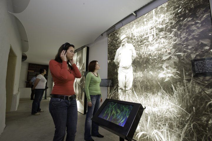 Zwei Frauen in einem Museum, die eine interaktive Ausstellung betrachten. Eine Frau trägt ein rotes Shirt und die andere ein grünes Langarm-Shirt. Ein Mann in einem weißen Outfit steht vor einem großen Bild.