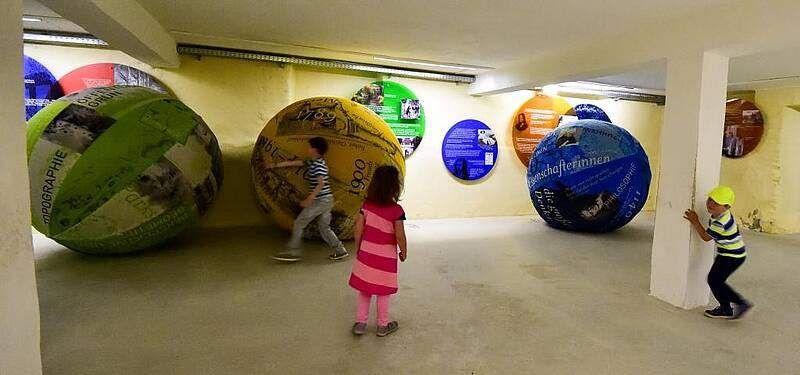 Drei Kinder spielen zwischen großen bunten Bällen in einem Raum. Ein Ball ist grün und weiß bemalt, ein anderer gelb und schwarz. Sie befinden sich in einer musealen Umgebung mit gelben Wänden.