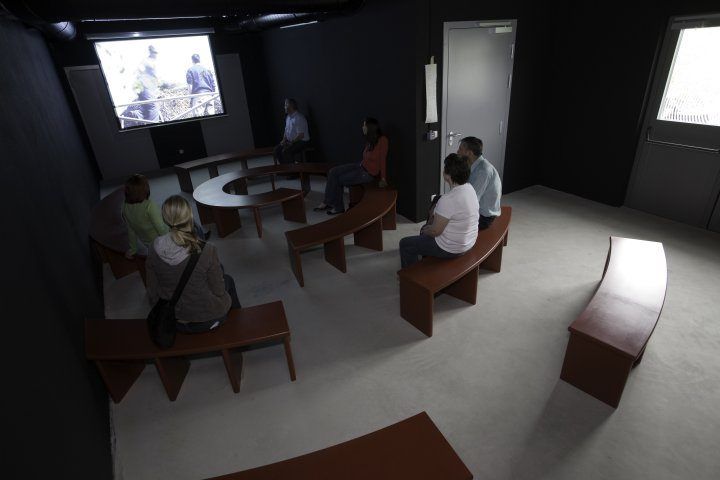 Eine Gruppe von Menschen sitzt auf Bänken in einem Raum und schaut sich ein Video auf einer Leinwand an. Der Raum ist dunkel mit einer Projektionsleinwand an der Wand.