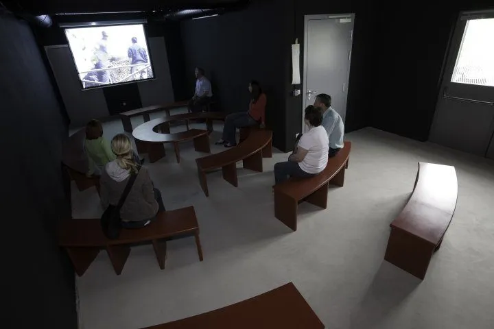 Eine Gruppe von Menschen sitzt auf Bänken in einem Raum und schaut sich ein Video auf einer Leinwand an. Der Raum ist dunkel mit einer Projektionsleinwand an der Wand.
