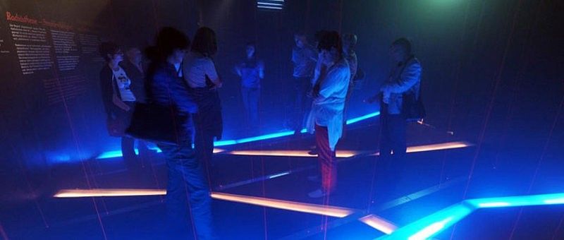 Eine Gruppe von Menschen steht in einem schwach beleuchteten Raum mit blauen Lichtern auf dem Boden, scheinbar Teil einer interaktiven Installation.