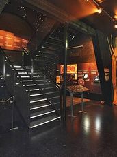 Eine moderne Treppe mit Glasstufen wird von Deckenleuchten beleuchtet. Der Bereich ist schwach beleuchtet, und es gibt ein Ausstellungsbrett und Poster an der Wand.