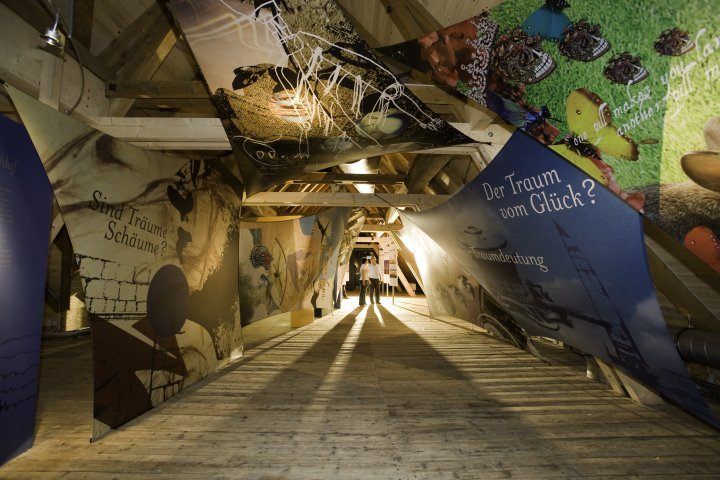 Ein Korridor mit Kunstinstallationen und deutschem Text. Zwei Personen gehen in der Mitte, umgeben von Licht.