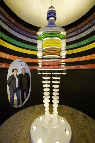 Eine bunte Plattenskulptur steht auf einer Holzplattform. Dahinter stehen zwei Menschen vor einem Spiegel, der eine Wand mit bunten Bändern reflektiert.