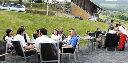 Eine Gruppe von Menschen in einem Restaurant am Hang sitzt um Tische herum, unterhält sich und genießt Getränke. Im Hintergrund ist ein Haus mit einer Steinmauer und mehreren geparkten Autos zu sehen.