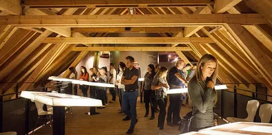 Eine Gruppe von Menschen erkundet eine Dachbodenausstellung mit Holzbalken und langen Tischen mit beleuchteten Objekten. Einige stehen und andere sitzen. Die Atmosphäre ist dunkel und intim.
