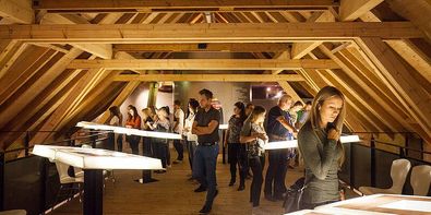 Eine Gruppe von Menschen erkundet eine Dachbodenausstellung mit Holzbalken und langen Tischen mit beleuchteten Objekten. Einige stehen und andere sitzen. Die Atmosphäre ist dunkel und intim.