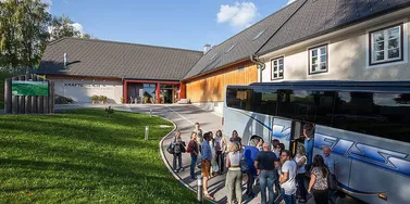 Eine Gruppe von Menschen steigt in einen Bus vor einem Gebäude mit der Aufschrift Kraftwerkreich. Das Gebäude hat ein geneigtes Dach und mehrere Fenster. Es gibt Bäume und Gras in der Umgebung.