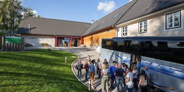 Eine Gruppe von Menschen steigt in einen Bus vor einem Gebäude mit der Aufschrift Kraftwerkreich. Das Gebäude hat ein geneigtes Dach und mehrere Fenster. Es gibt Bäume und Gras in der Umgebung.