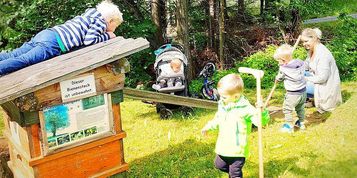 Eine Familie mit drei Kindern ist draußen. Das älteste Kind klettert auf eine Holzstruktur, während das Baby in einem Kinderwagen sitzt. Die Mutter hält ein weiteres Kind. Sie sind auf einer Rasenfläche.