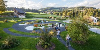 Luftaufnahme eines Parks mit spazierengehenden Personen. Der Park bietet verschiedene Pflanzen und einen geschwungenen Weg. Im Hintergrund befinden sich Häuser und Bäume.