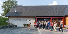 Eine Gruppe von Menschen steht vor einem Gebäude mit der Aufschrift 'Krafte Reich'. Ein Mann in einer karierten Shorts führt sie an. Topfpflanzen und eine Steinmauer sind in der Nähe.