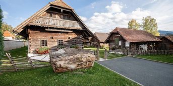 Ein traditionelles Holzhaus mit einem großen Baumstamm davor, in einem Dorf mit Garten, umgeben von Zäunen und einem Schild.