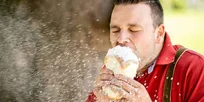Ein Mann in einem roten Hemd und mit Trägerhosen isst einen mit Puder überzogenen Kuchen im Freien, wobei Pulver herumfliegt.