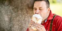 Ein Mann in einem roten Hemd und mit Trägerhosen isst einen mit Puder überzogenen Kuchen im Freien, wobei Pulver herumfliegt.