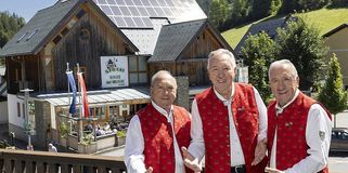 Drei ältere Männer stehen lächelnd auf einem Balkon vor einem Gebäude mit Solarmodulen. Ein Schild über ihnen sagt 'Stoane Haus der Musik.' Bäume, Pflanzen und eine Flagge sind in der Nähe. Im Hintergrund ist ein geparktes Auto zu sehen.