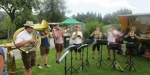 Eine Gruppe von Menschen spielt Instrumente auf einem Grasfeld. Sie tragen Hüte, Uhren und Turnschuhe. Einige von ihnen halten Blechblasinstrumente, während andere Trompeten halten. Hinter ihnen stehen Notenständer mit Papieren darauf. In der Ferne befindet sich ein grünes Zelt, ein Tisch und ein Mülleimer.