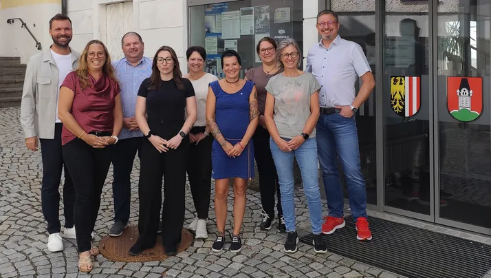 Eine Gruppe von Menschen steht vor einem Gebäude, lächelt und posiert für ein Foto. Sie sind lässig gekleidet und scheinen Kollegen zu sein.