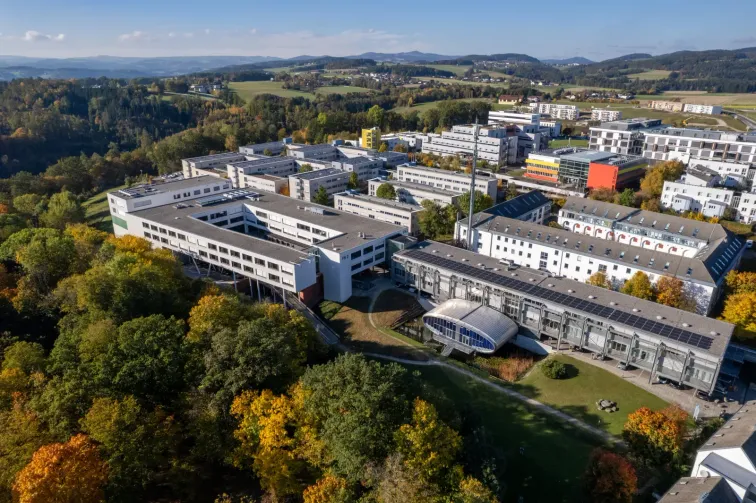Ein Luftbild eines modernen Campus mit mehreren Gebäuden, umgeben von üppigem Grün und Bäumen. Der Campus liegt vor einer Kulisse aus sanften Hügeln und einem klaren blauen Himmel.