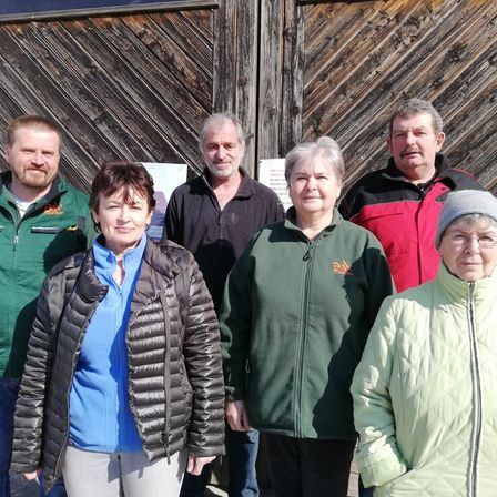Sechs Personen posieren für ein Foto vor einer Holztür. Ein Mann trägt eine grüne Jacke, eine Frau ein blaues Shirt, eine weitere Frau eine grüne Jacke, ein weiterer Mann eine rot-schwarze Jacke, ein weiterer Mann eine grüne Jacke und die letzte Frau eine hellgrüne Jacke.