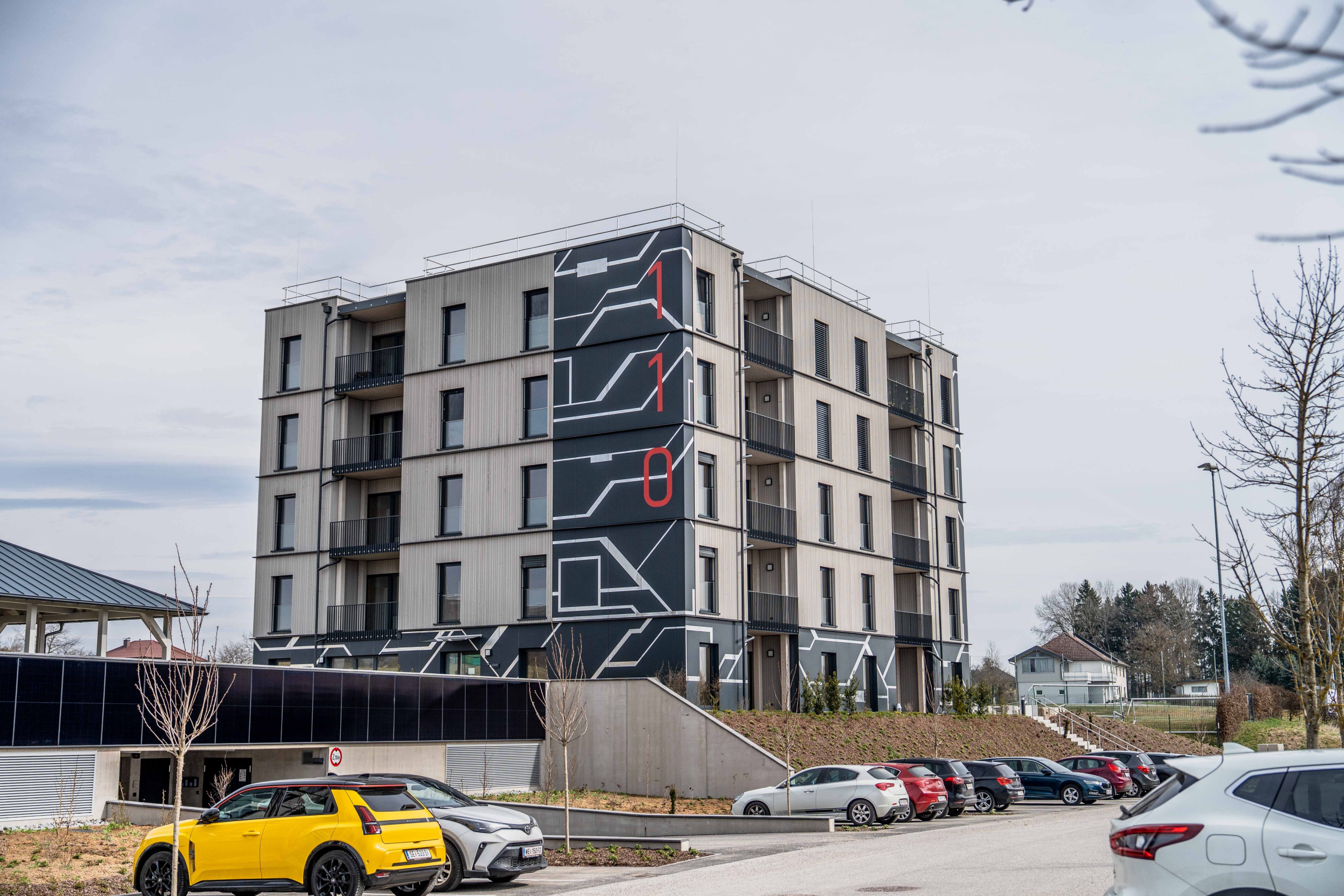 Ein modernes Wohngebäude mit geometrischem Design an der Fassade, das die Zahlen 110 in Rot zeigt. Das Gebäude hat mehrere Stockwerke mit Balkonen und ist von geparkten Autos umgeben.