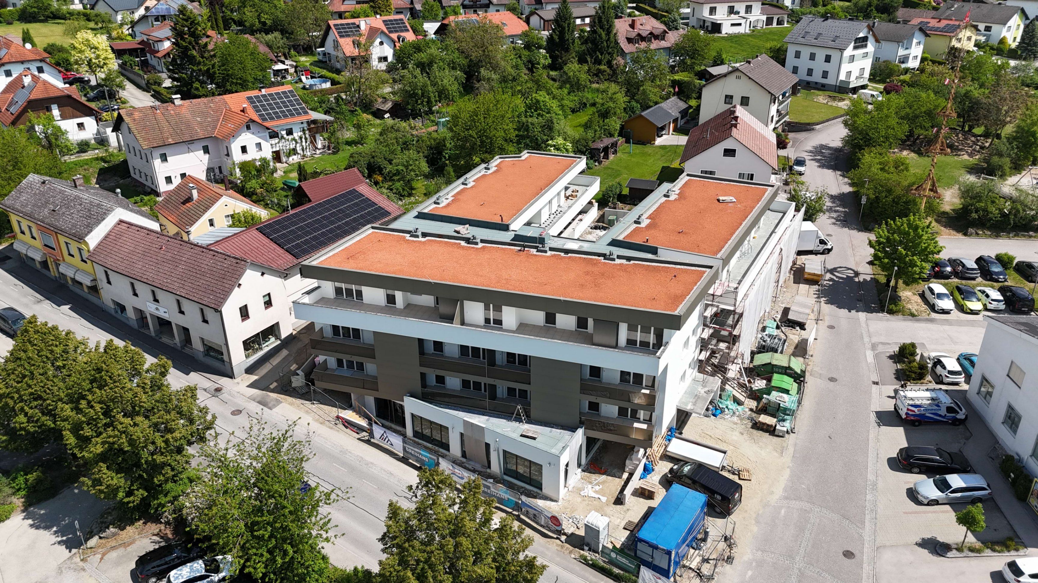 Ein Luftbild eines im Bau befindlichen Gebäudes mit roten Dächern und Solarmodulen, umgeben von Häusern, Bäumen und einer Straße.