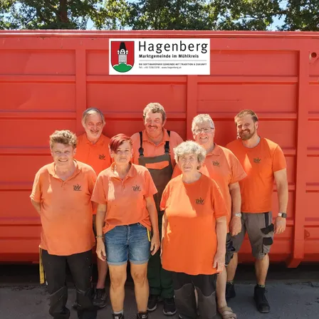 Sieben Menschen in orangefarbenen Shirts stehen vor einem roten Gebäude. Sie lächeln und posieren für ein Foto. Das Gebäude hat ein Schild mit dem Wort 'Hagenberg' darauf.