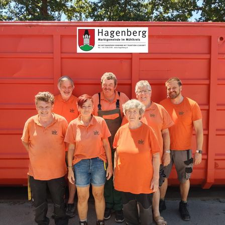 Sieben Menschen in orangefarbenen Shirts stehen vor einem roten Gebäude. Sie lächeln und posieren für ein Foto. Das Gebäude hat ein Schild mit dem Wort 'Hagenberg' darauf.