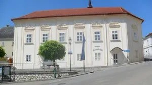Ein großes weißes Gebäude mit rotem Dach und vielen Fenstern, an einer Straße mit einem Baum davor.