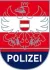 Das Wappen der österreichischen Polizei, mit einem gekrönten Adler mit ausgebreiteten Flügeln, der ein Schwert und einen Schild hält, auf einem roten und weißen Hintergrund mit dem Wort 'POLIZEI' darunter in fetter Schrift.