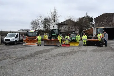 Eine Gruppe von Menschen in gelben Sicherheitsanzügen steht vor einer Reihe von Schneepflügen und Fahrzeugen.