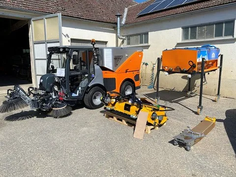 Ein orange-graues Fahrzeug mit einer großen Bürstenaufsatz ist vor einem Gebäude mit Solarpanelen auf dem Dach geparkt.