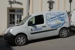 Ein weißer Lieferwagen mit blauem Logo und Text 'Wasser für Leben' steht vor einem Gebäude mit Glastür und Fenstern.