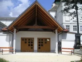 Ein weißes Gebäude mit einem braunen Dach und zwei Holztüren. Vor dem Gebäude befinden sich zwei Bänke.