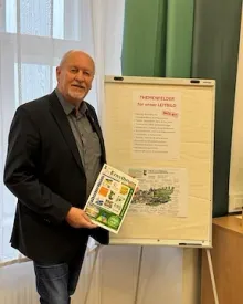 Ein älterer Mann in einem schwarzen Anzug und einem grauen Hemd steht neben einem Whiteboard mit einem Poster darauf. Er hält ein Buch mit dem Titel 'Ernstern'.