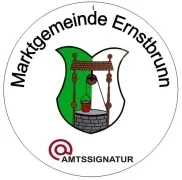 Ein rundes Emblem zeigt einen Brunnen mit einem Eimer, umgeben von einem grünen Blatt und einer Ziegelmauer. Der Text 'Marktgemeinde Ernstbrunn' ist in einer geschwungenen Form um das Emblem herum geschrieben. Unten steht der Text 'AMTSIGNATUR' in einer roten, stilisierten Schrift.