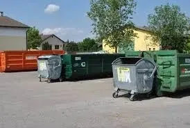 Ein Parkplatz mit mehreren Müllcontainern, darunter ein grüner mit einem weißen Etikett, ein grauer und ein orangefarbener. Hinter den Containern befinden sich Gebäude, von denen eines eine gelbe Wand hat.