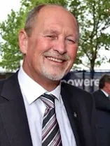 Ein älterer Mann mit weißem Bart und Schnurrbart lächelt und schaut in die Kamera. Er trägt einen schwarzen Anzug, ein weißes Hemd und eine gestreifte Krawatte.