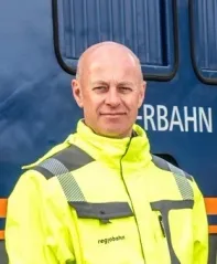 Ein Mann in einem gelben Sicherheitsjacke mit dem Wort 'Regiobahn' darauf steht vor einem blauen Zug.