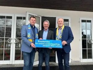 Drei Männer stehen vor einem Gebäude und halten ein blaues Banner mit weißen Texten. Der Mann auf der linken Seite trägt einen blauen Mantel mit einem gelben und blauen Schal. Der Mann in der Mitte trägt einen schwarzen Anzug. Der Mann auf der rechten Seite trägt einen blauen Mantel mit einem gelben Schal.